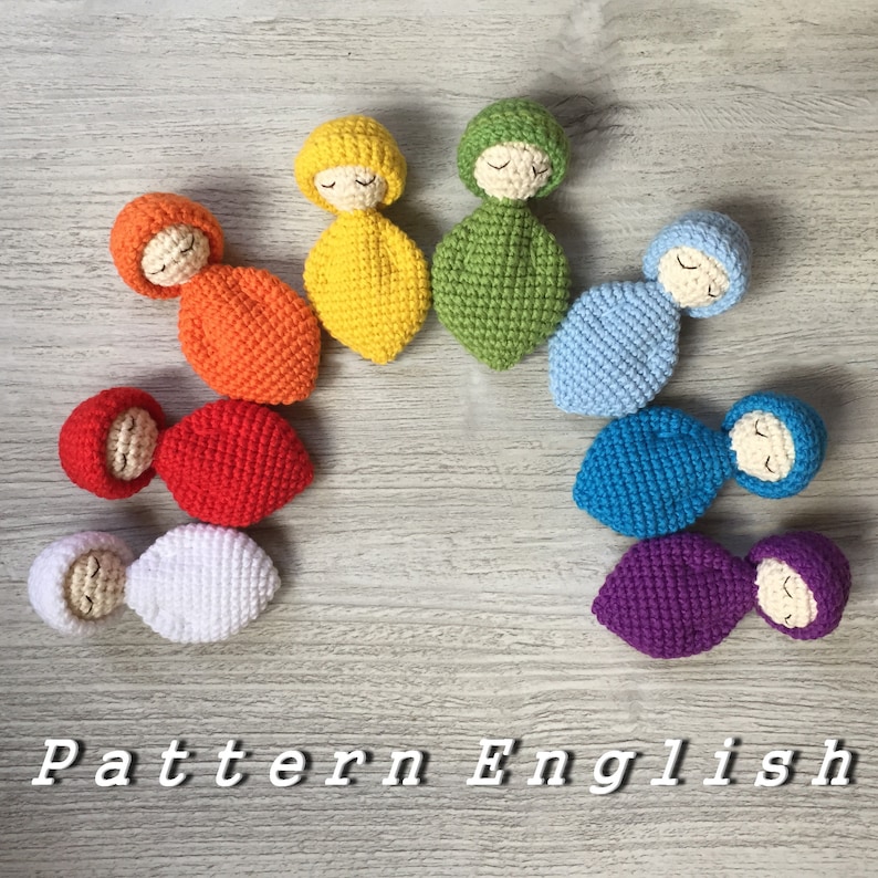 May include: A rainbow of crocheted baby dolls, each with a different color and a sleeping face. The dolls are arranged in a semi-circle, with the yellow doll at the top and the purple doll at the bottom.