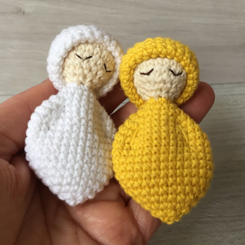 May include: Two crocheted dolls, one white and one yellow, with closed eyes. The dolls are wearing simple crocheted outfits.