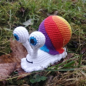 Puede incluir: Un juguete de ganchillo en forma de caracol con una concha de arcoíris y un cuerpo blanco. El caracol tiene ojos grandes y blancos con pupilas negras y una pequeña boca negra.