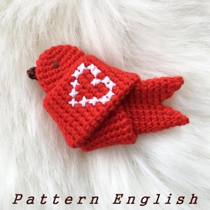 May include: Red crocheted bird with a white heart on its chest. The bird is lying on a white fluffy surface. Pattern English.