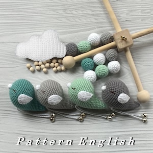 Puede incluir: Un móvil para bebé de crochet con cuatro pájaros grises y verdes, una nube blanca y cuentas de madera. El móvil está colgado de un marco de madera.
