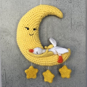 May include: A crocheted yellow crescent moon with a smiling face and a white crocheted goose sleeping inside. Three yellow crocheted stars hang from the moon.