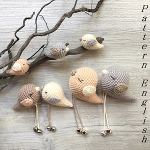 May include: Five crocheted bird ornaments with bells hanging from a branch. The birds are in shades of beige, brown, and grey.
