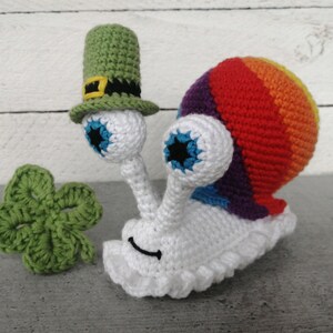 以下が含まれることがあります： 虹色の貝殻と大きな青い目をした白い編みぐるみのカタツムリ。カタツムリは、黒いリボンと黄色のバックルが付いた緑色の編みぐるみの帽子をかぶっています。手前に緑色の編みぐるみの四つ葉のクローバーがあります。