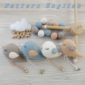 Pode incluir: Um móbile para bebê com uma nuvem branca, três pássaros de crochê em azul, branco e bege e contas de madeira. Os pássaros têm pequenos sinos presos aos pés. O móbile está pendurado em uma estrutura de madeira.