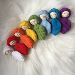 May include: A set of ten crocheted dolls in rainbow colors, each with a closed-eye expression and a small, rounded hat. The dolls are arranged in a semi-circle on a white fluffy surface.