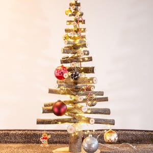 Könnte beinhalten: Ein dekorativer Weihnachtsbaum aus gestapelten horizontalen Holzästen auf einem runden Holzsockel. Der Baum ist mit warmweißen Lichterketten, roten und goldenen Weihnachtskugeln, einem silbernen Ornament, einem Lebkuchenmann und Zuckerstangen geschmückt.