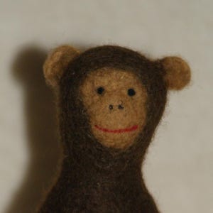 Può includere: Una figura di scimmia in feltro fatta a mano con un corpo marrone e un viso, orecchie e bocca cucita in rosso marrone chiaro. La scimmia è posta sopra un portauovo di legno chiaro, creando un oggetto decorativo stravagante.