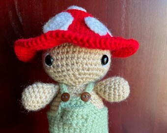 May include: A crocheted mushroom-shaped toy with a red and white hat, brown eyes, and green overalls.