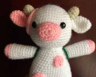 May include: White crocheted stuffed cow with pink ears, nose, and spots. The cow has black eyes and a green strap around its back.