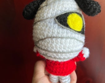 May include: A crocheted stuffed animal resembling a mummy with a black nose and ears, a yellow eye, and a red shirt.