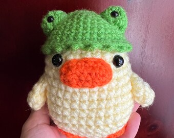 May include: A crocheted yellow duck wearing a green frog hat with black eyes.