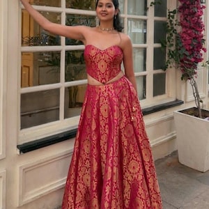 May include: A two-piece formal outfit in a vibrant fuchsia hue with gold floral patterns. The top is a strapless crop top, and the skirt is a flowing, floor-length design. The outfit is suitable for special occasions.