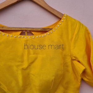 May include: A yellow blouse with a round neckline and short sleeves. The neckline is decorated with a row of white pearls. The blouse has a keyhole opening in the back.