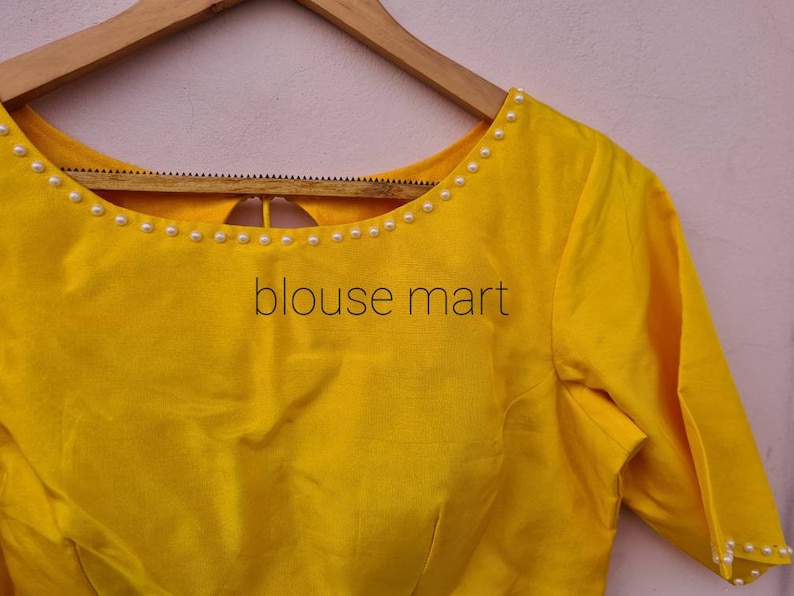 May include: A yellow blouse with a round neckline and short sleeves. The neckline is decorated with a row of white pearls. The blouse has a keyhole opening in the back.