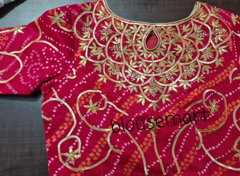May include: A red blouse with a gold and white embroidered design. The embroidery features a floral pattern with leaves and beads. The blouse has a round neckline and a keyhole opening at the back.