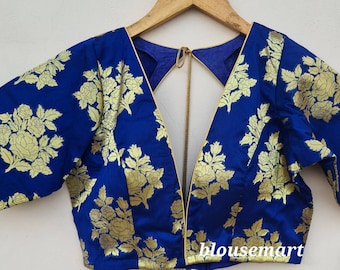Peut inclure: Un chemisier bleu en brocart avec un décolleté en V profond et un dos à ouverture. Le chemisier présente un motif floral doré et une bordure dorée autour du col et des manches. Le chemisier est sur un cintre.