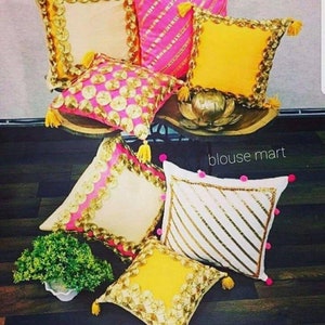 May include: A set of six decorative throw pillows with gold and pink embellishments. The pillows are made of a variety of fabrics, including velvet, satin, and cotton. The pillows are arranged on a wooden surface with a green plant in the foreground. The text "blouse mart" is visible in the background.