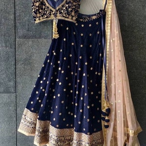 Puede incluir: Un conjunto lehenga de terciopelo azul marino con bordados florales dorados. El conjunto incluye un top sin mangas y una falda acampanada con un borde dorado. Un dupatta melocot&oacute;n transparente con detalles dorados completa el conjunto. El atuendo se exhibe en una percha de madera.