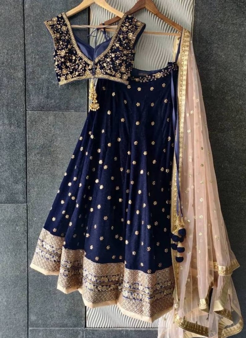 Puede incluir: Un conjunto lehenga de terciopelo azul marino con bordados florales dorados. El conjunto incluye un top sin mangas y una falda acampanada con un borde dorado. Un dupatta melocot&oacute;n transparente con detalles dorados completa el conjunto. El atuendo se exhibe en una percha de madera.