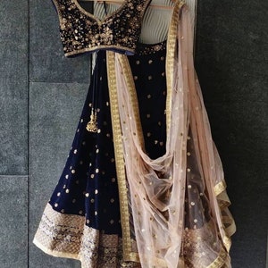 Puede incluir: Un conjunto lehenga de terciopelo azul marino con bordados y adornos dorados. El conjunto incluye un top sin mangas, una falda larga y una dupatta transparente de color rosa p&aacute;lido con detalles dorados. El atuendo est&aacute; colgado en una percha de madera.