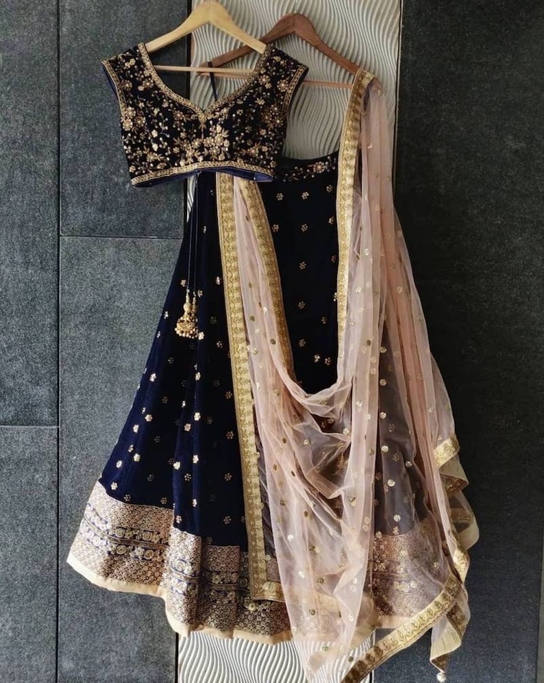 Puede incluir: Un conjunto lehenga de terciopelo azul marino con bordados y adornos dorados. El conjunto incluye un top sin mangas, una falda larga y una dupatta transparente de color rosa p&aacute;lido con detalles dorados. El atuendo est&aacute; colgado en una percha de madera.