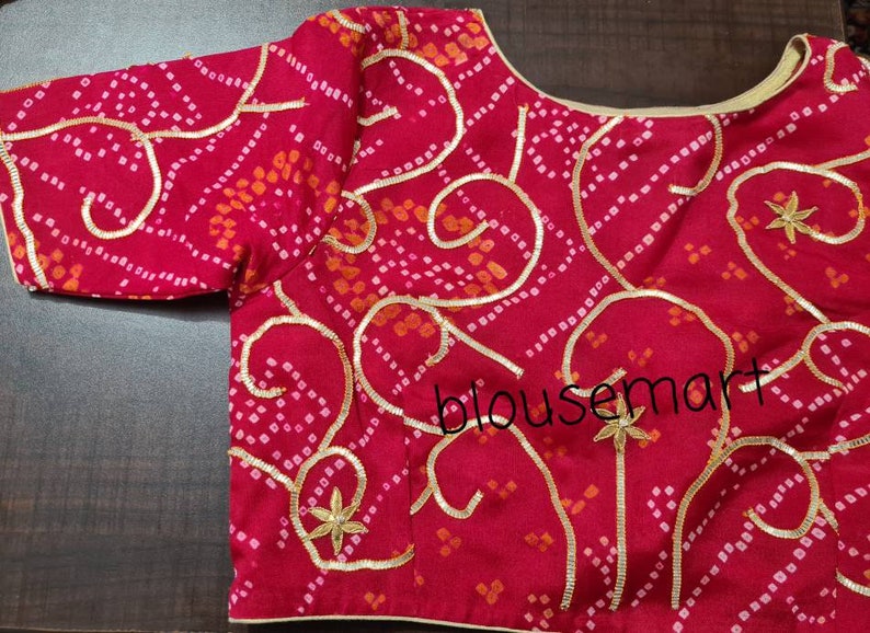 May include: A red and white patterned blouse with gold embroidery and a scoop neckline. The blouse has a "blousemart" watermark.