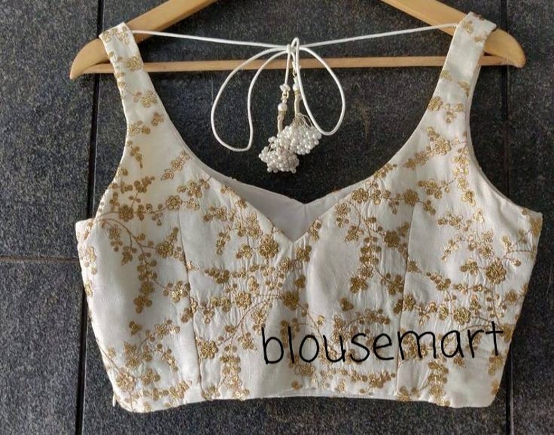 May include: White sleeveless blouse with a gold floral pattern and a white tassel tie. The blouse has a scoop neckline and is embroidered with gold thread. The text "blousemart" is visible in the bottom right corner of the image.
