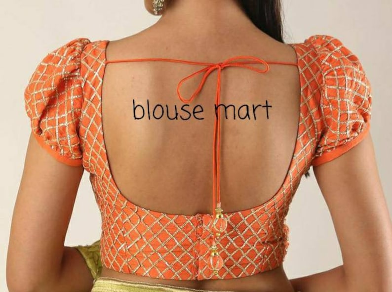 May include: Orange blouse with a low back and puff sleeves. The blouse features a diamond pattern and is decorated with gold embroidery. The blouse has a tie at the back.