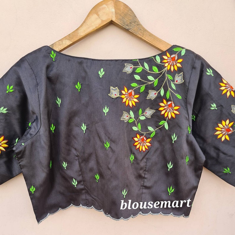 Op de afbeelding: Een zwarte zijden blouse met een gegolfde zoom en een geborduurd bloemenontwerp in tinten groen, geel, oranje en grijs. De blouse heeft een boot hals en korte mouwen. De tekst "blousemart" is zichtbaar onderaan de afbeelding.