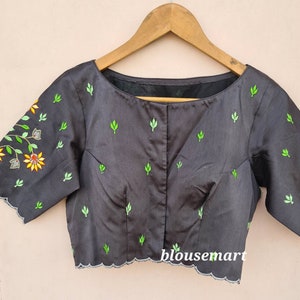 Op de afbeelding: Zwarte blouse met groene en gele bloemenborduursel. De blouse heeft een ronde hals en korte mouwen. Het borduursel bevindt zich op de voorkant en de mouwen van de blouse. De blouse heeft een gegolfde zoom.