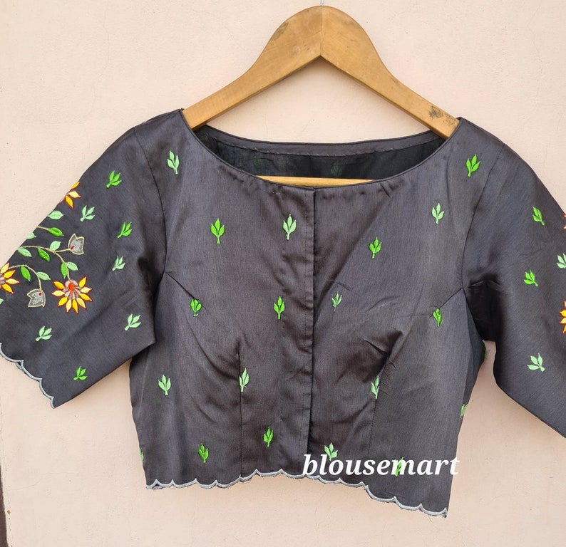 Op de afbeelding: Zwarte blouse met groene en gele bloemenborduursel. De blouse heeft een ronde hals en korte mouwen. Het borduursel bevindt zich op de voorkant en de mouwen van de blouse. De blouse heeft een gegolfde zoom.