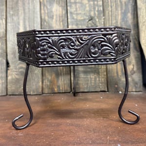 May include: A dark brown metal decorative stand with an ornate, floral design. The stand has a rectangular top and curved legs. The stand is approximately 6 inches long, 4 inches wide, and 4 inches tall.