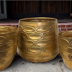 May include: Three gold-colored metal planters with an oval pattern. The planters are different sizes and sit on a concrete surface. The largest planter is approximately 18 inches in diameter and 16 inches tall. The planters are empty.