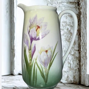 May include: A tall, white ceramic pitcher with a curved handle and spout. The pitcher is decorated with a floral design featuring purple and white crocus flowers with green stems and leaves. The background has a gradient of green at the bottom.