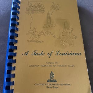 May include: A gold-colored spiral-bound cookbook titled A Taste of Louisiana, compiled by the Louisiana Federation of Women's Clubs. The cover features line drawings of a building, a pelican, and a church, with Claitor's Publishing Division text.