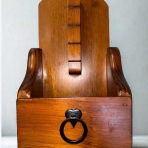May include: A wall-mounted wooden storage box with a medium brown finish. The unit features a tall, arched back panel with a vertical wooden slat and a front compartment adorned with a black metal ring pull handle shaped like a heart.