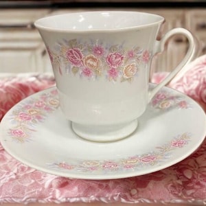 May include: A white teacup and saucer set with a floral design in pink, peach, and green. The teacup has a curved handle and sits on a matching saucer. The floral pattern is repeated on both pieces, creating a cohesive look.