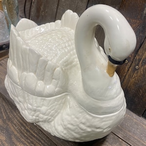 May include: A white ceramic swan shaped serving dish with a lid. The swan's head is turned to the side and the lid is closed.