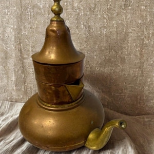 May include: A vintage copper teapot with a bulbous base, a cylindrical midsection, and a conical lid topped with a decorative finial. The spout is curved and golden. The teapot is likely used for brewing tea or coffee.