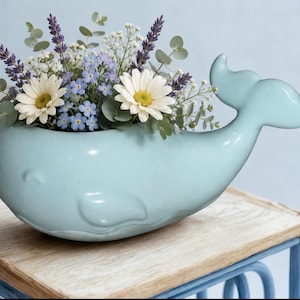 May include: A light blue ceramic whale-shaped planter filled with a floral arrangement. The arrangement includes white daisies, blue forget-me-nots, lavender, and greenery. The planter sits on a wooden table.
