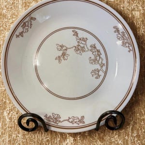 May include: White ceramic plate with a brown floral design and a brown border. The plate is displayed on a black metal stand. The floral design is a branch with flowers. The plate is round.