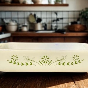 May include: A rectangular, off-white Pyrex baking dish with a floral design in green. The dish is empty and sits on a wooden surface. The kitchen background includes a stove, shelves, and various kitchen items.