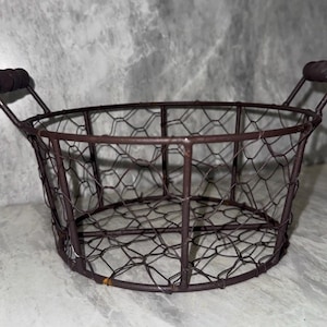 May include: A round, dark brown wire basket featuring a hexagonal chicken wire mesh pattern. The basket has a sturdy metal frame and two small, cylindrical dark brown wooden handles attached to the sides for easy lifting and carrying.