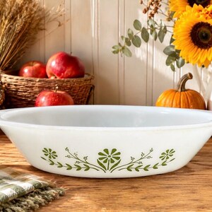 May include: A white oval baking dish featuring a green floral pattern along the exterior side. The dish has a smooth, glossy finish with a wide, flat rim and a shallow, rounded basin, designed for oven use and serving various food items.