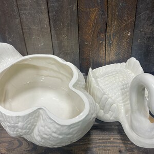May include: A white ceramic swan-shaped serving dish with a removable lid. The dish is designed to look like a swan with its head turned to the side. The lid is shaped like the swan's back and wings.