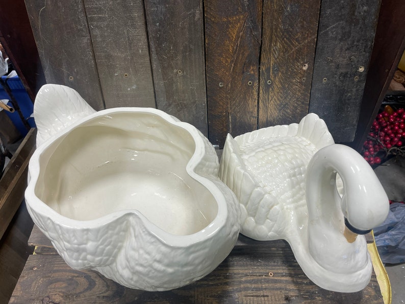 May include: A white ceramic swan-shaped serving dish with a removable lid. The dish is designed to look like a swan with its head turned to the side. The lid is shaped like the swan's back and wings.