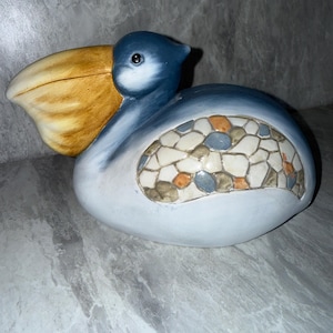 May include: A ceramic pelican figurine featuring a large yellow beak, a blue head and body, and a decorative wing panel composed of a mosaic pattern with white, blue, and orange stone-like segments. The bird is depicted in a resting position.