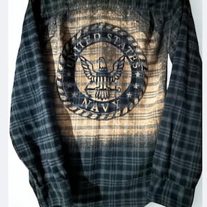 May include: Black and gray plaid flannel shirt with a distressed United States Navy emblem on the back. The emblem features an eagle, stars, and the word "NAVY" in a circular design.