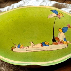 May include: A green ceramic serving dish with a whimsical beach scene painted on the interior. The scene depicts figures relaxing under a palm tree. The dish has a scalloped edge with a cream-colored border.